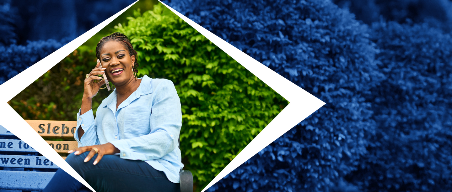 An African American woman wearing a light blue button up blouse sits on a bench. She is holding her cell phone to her right ear with her right hand, and is smiling while on the phone. Behind her is all greenery from shrubs or bushes.