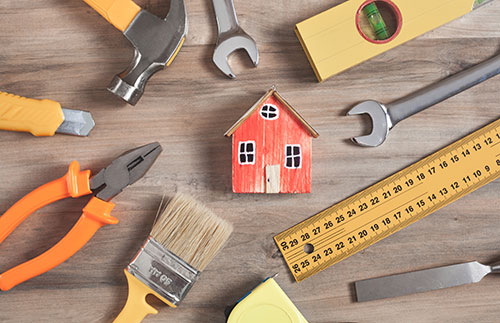 Home Improvement tools on a wooden table surrounding a mini wooden house.