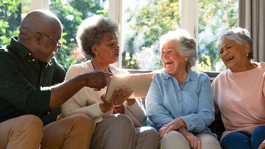 A group of four senior citizens sitting on a couch together and laughing while using an iPad.