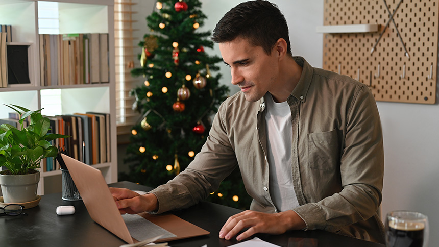A man sitting next to a Christmas tree in his office trying to avoid E-Card Scams on his laptop.