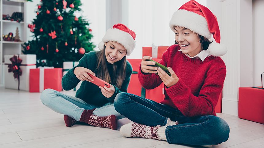 Children wearing Santa hats and sitting next to a Christmas tree while they play games on their phones.