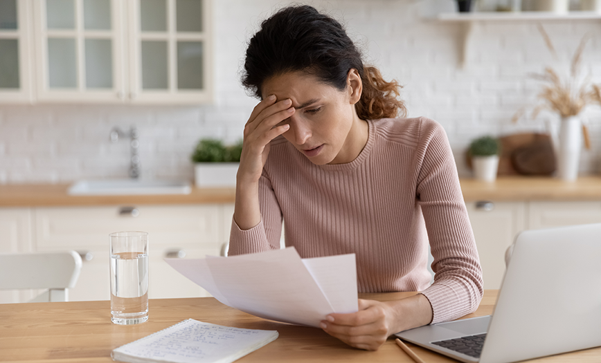 A woman is sitting in her kitchen. She appears frustrated. She is holding papers in her left hand and is looking down at them, her right hand is on her forehead. She is wearing a very light pink long sleeve shirt. To her left, is an opened silver laptop. This image represents the blog, "FBI Issues Alert on Discount Medical Insurance Scams."