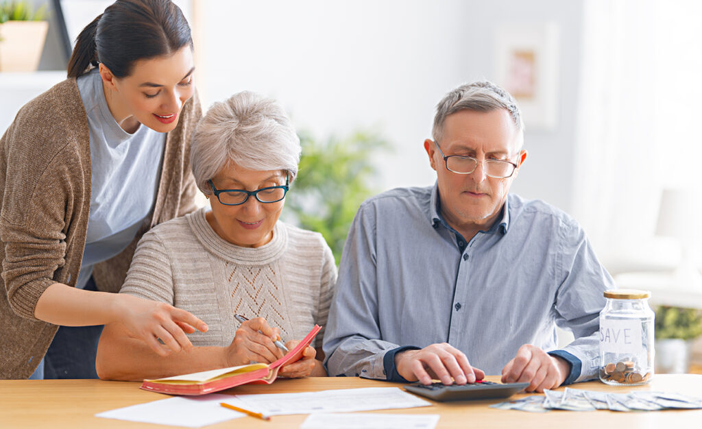 older couple sitting at a table with papers, a calculator and note book with a woman standing behind them. showing how to help aging parents with finances.