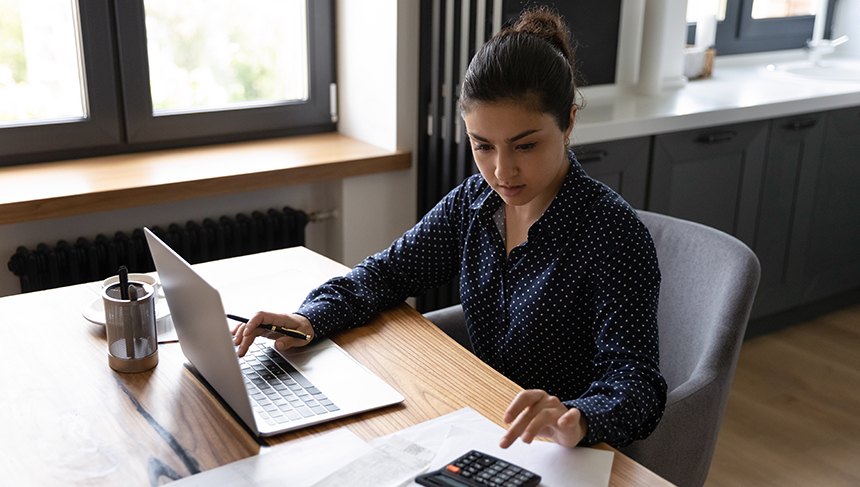 This image goes with the blog, "Beginner's Guide to Managing Finances for Small Businesses." A woman sits in a grey office chair at a desk. A silver laptop is open, her right hand hovers over the keyboard while holding a black and gold pen. She looks down to her left at a calculator. Her left hand is overing over the calculator like she is going to press buttons.