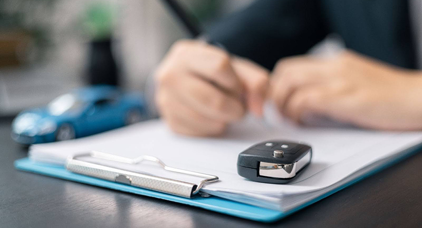 A blue clipboard is on a table with papers clipped on it. There is a car key fob on the top left of the papers. There are two hands close together on top. The right hand has a pen and it looks like the person is signing the paper. This image is for the blog, Answering, "Can I Get a Car Loan With a New Job?"