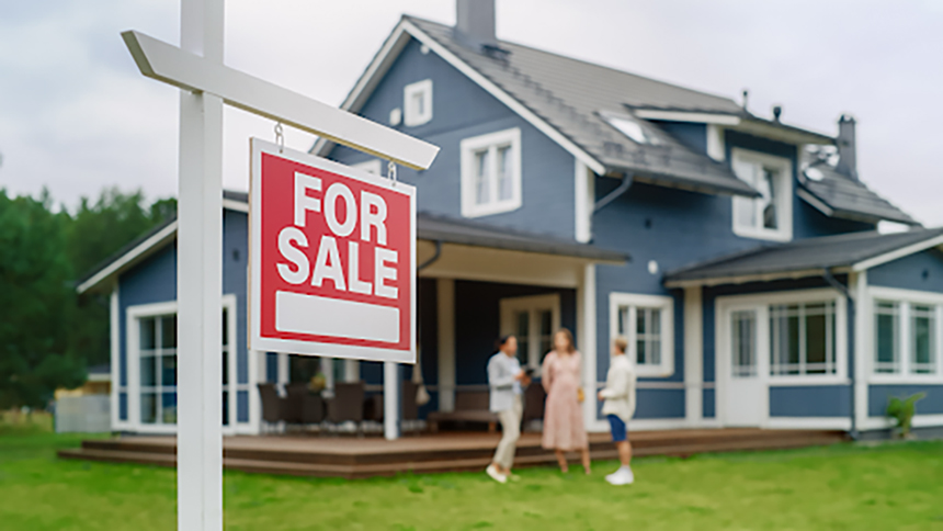This image is used to represent the myths about buying a home. We see an image of a dark blue house that is out of focus in the background with a man and woman talking to their realtor. In the front left of the image is a red for sale sign hanging on a white post.