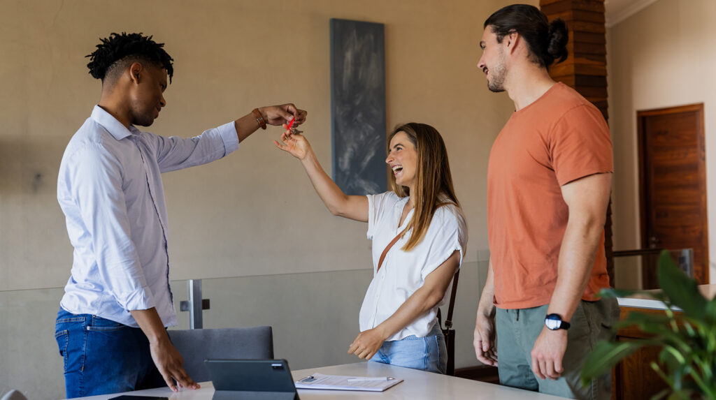 A realtor on the left is handing the keys to his clients who are a woman and man that appear to be in their late 20s or early 30s.