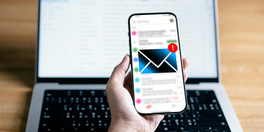 A laptop with a bright white screen is in the background and a hand holiding a cell phone is in the foreground. On the cell phone screen is an envelope icon and a red alert above it.