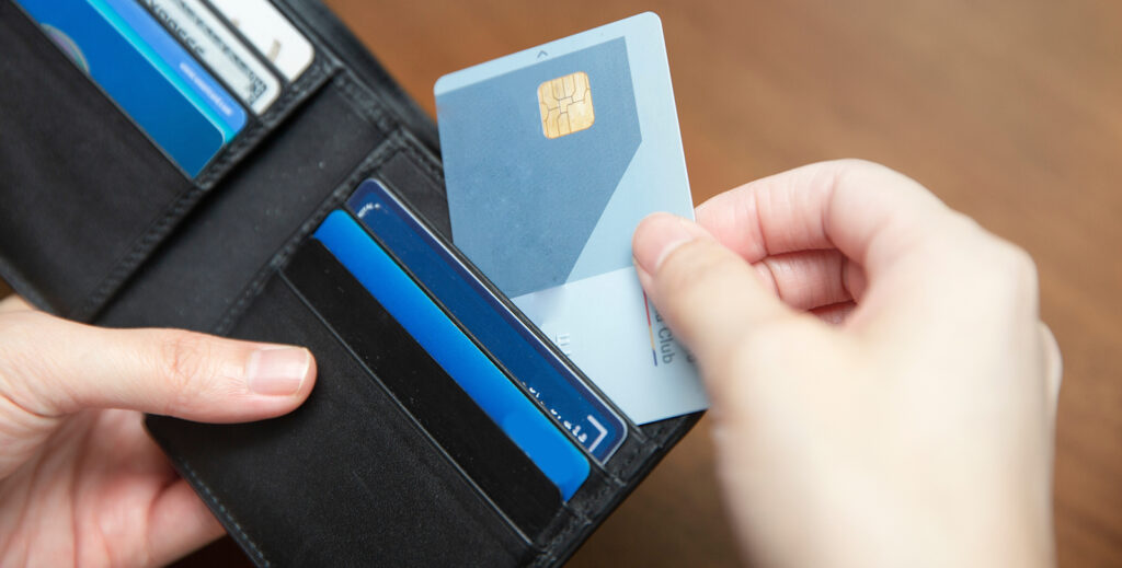 Someone is holding a black wallet open with their left hand. There are 7 credit cards that fill the slots of the wallet. The right hand is taking one of the cards out of the wallet.