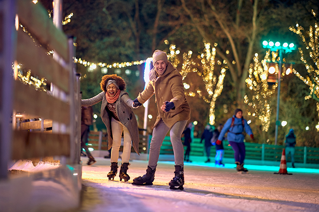 A man and a woman holding hands while ice skating in a park with warm white Christmas lights on the trees. This image represents affordable holiday fun.