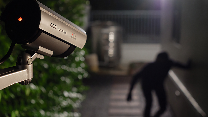 This image shows home security cameras monitoring a criminal. It's dark outside. In the top left of the image, a camera is coming out pointing towards the bottom right. There is a person completely shadowed out dressed in all black, creeping up the side of a building on the right.