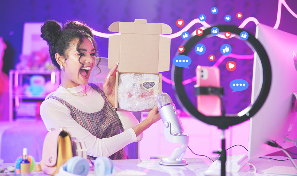 woman social media influencer laughing while holding a box towards her phone recording a video with like, love, and comment social media icons around.