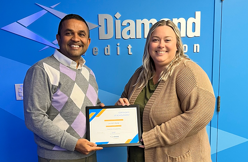 Diamond Credit Union's President/CEO, Rick Patel (left), handing Diamond Credit Union's Director of Payment and Operational Services, Lauren Harp (right), her certificate for completing Diamond's Essentials of Leadership course in October 2024. They are posed in front of a blue wall with the Diamond logo directly behind them.