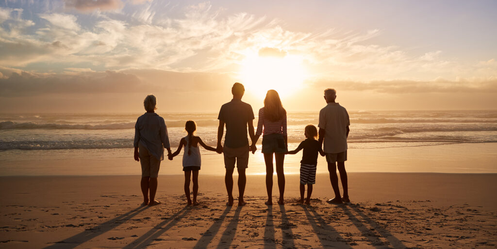 A multi generational family is pictured standing on a beach in front of the ocean. There is a grandma on the left, then a small child, a father, mother, another child and then a grandfather. The photo represents the sandwich generation and caring for children and parents at the same time.