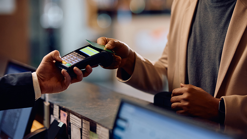This image shows a person using there card to tap to pay at a point of sale. There is a hand from the left of the image holding a point of sale system out to the customer. The customer is on the right side and has their credit card on top of the point of sale. This image is for the article about new NFC scams that can give your card information away with a tap.