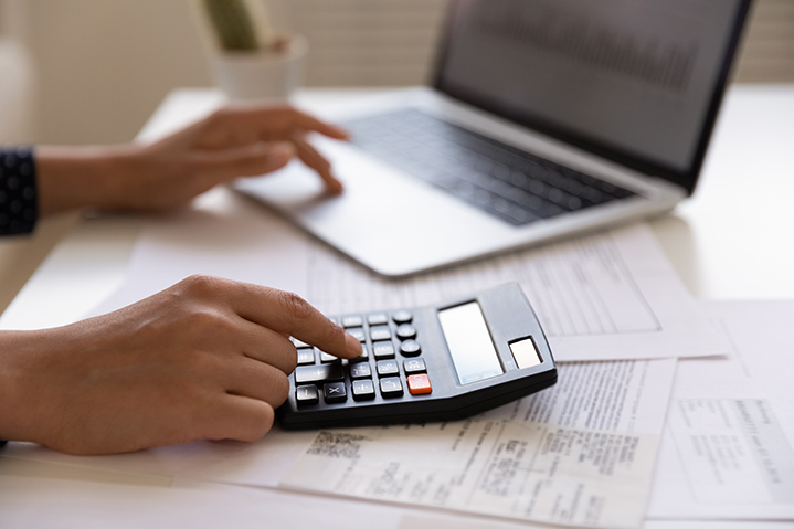 An image to represent someone preparing for a business loan. there is a desk with a laptop in the background and two hands are leaning on the desk from the left side. One hand is one the laptop scrolling the mouse, and the right hand is typing on a calculator.