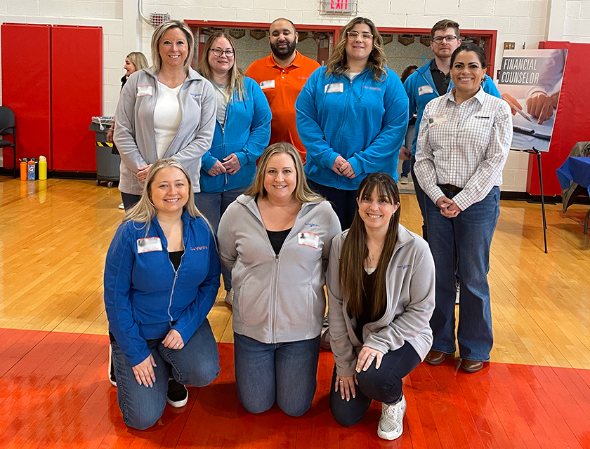 This photo shows Diamond Credit Union volunteers inside the gym of Owen J. Roberts High School at one of the Financial Reality Fairs they participated in for the 2024-2025 school year. There are nine people in total, five people are standing in the back and three women are kneeling in front. Behind them is a sign that says. "Financial Counselor," which is one of the stations at the fair.