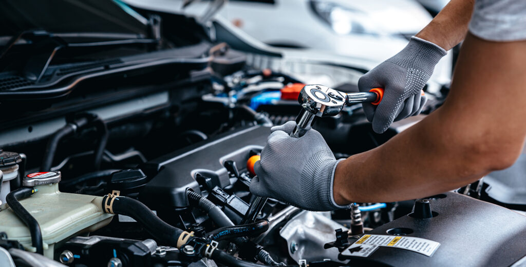 This image represents the blog, "The Pros & Cons of Auto Warranties." Two arms are reaching into a car with it's hood up and engine exposed, doing some kind of repair work.