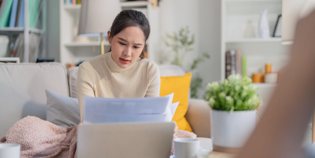 A woman in here 20s sits on her couch and is looking at her laptop as well as some other papers. The image is to represent a person going over their finances to build financial confidence.