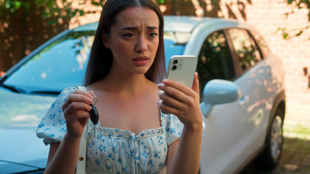 A woman is in the foreground of the photo with a white car in the background. She's holding her keys up in her right hand, and has her cell phone in her left hand. She looks distressed. This photo represents someone who entered an upside-down auto loan.
