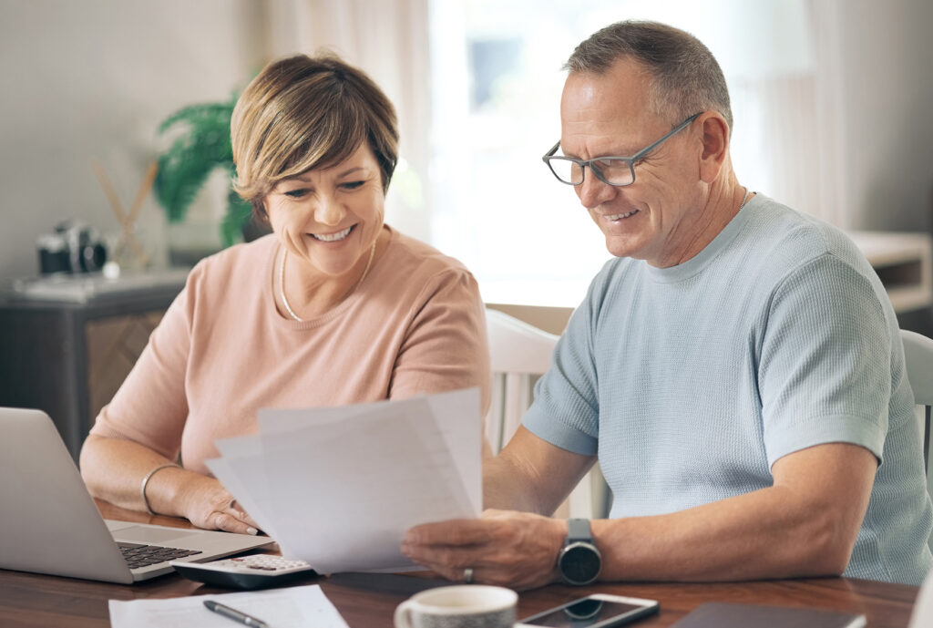 a couple sitting at a kitchen table with a laptop. man holding paper and woman looking at it completing their yearly financial check-in.