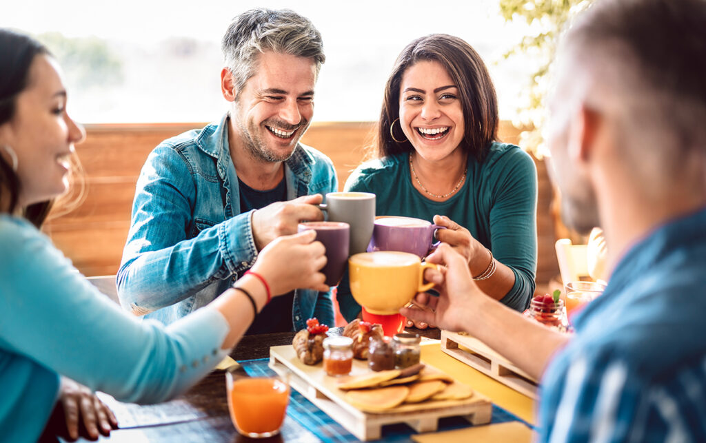 group of friends smiling and laughing clinking mugs while enjoying coffee and desserts at a resaturant.