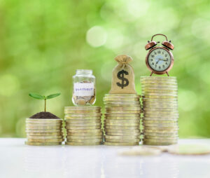 share certificates representation. green bokeh background with 4 stacks of various height coins. on top of the stacks is a sprout out of dirt, clear jar with coins and the word investment, sack of money, and alarm clock.