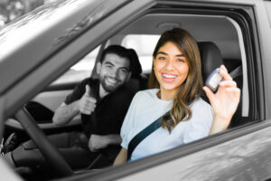 young female and male sitting in a car. male in the passenger seat is smiling with a thumbs up and the female is smiling holding up a key showing she is buying a car.