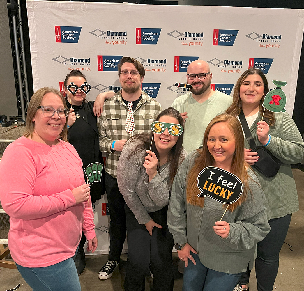 Seven attendees of Diamond Credit Union's All in for Hope, posed for a picture in front of a photo wall that has the American Cancer Society logo and Diamond Credit Union's logo on it. 