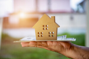 loan promotions image. person's hand holding money bills with a wooden house on top.