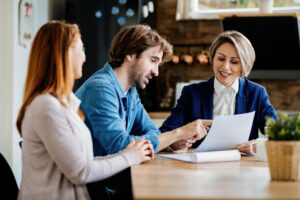 man and woman sitting at a table with a female professional. Professional is holding a paper and man is pointing at it while the woman is looking at it and smiling representing a loan review.