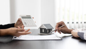 table in an office with a house figurine and contract. Left has a person's hands holding a calculator and right side has a person's hand holding a pen ready to sign the contract.