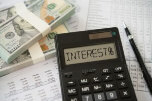 image of 100 bill straps laying on top of financial papers. on top of the papers is a calculator with the words "INTEREST%" and a pen.