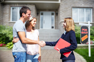 man and woman standing in front of a house shaking hands with a female realtor going through the house hunting process