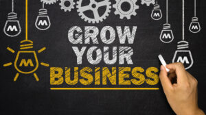 business loans image. hand holding a piece of chalk. Behind is a chalkboard with white and yellow chalk drawings of light bulbs and cog wheel. Text says "GROW YOUR BUSINESS".