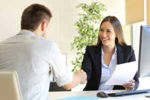 Business woman sitting in front of a computer holding a paper and shaking the hand of a man sitting across from her.