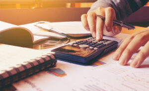 person holding a pen and typing on a calculator on a table with papers and notebooks around portraying a person using our financial calculators.